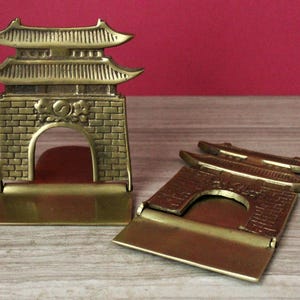 May include: A pair of brass bookends featuring a traditional East Asian gate design with a brick-patterned base, a central arched opening, and a multi-tiered roof structure. One bookend stands upright while the other is folded flat.