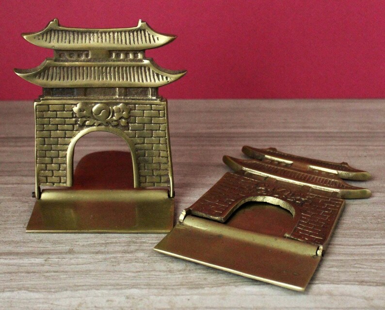 May include: A pair of brass bookends featuring a traditional East Asian gate design with a brick-patterned base, a central arched opening, and a multi-tiered roof structure. One bookend stands upright while the other is folded flat.