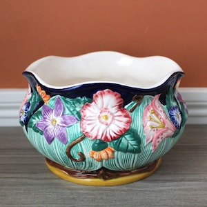 May include: A ceramic bowl featuring a scalloped rim and a deep blue interior. The exterior is decorated with raised, colourful floral reliefs including purple, pink, and orange flowers set against a textured green leaf background on a yellow base.