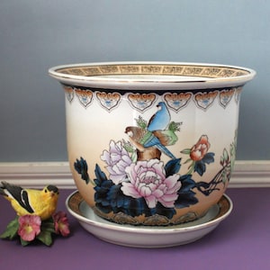 May include: A ceramic planter with a matching saucer featuring a cream-colored gradient background, painted pink peonies, dark blue leaves, and two birds perched on a branch. The rim has a decorative gold and black geometric border pattern.