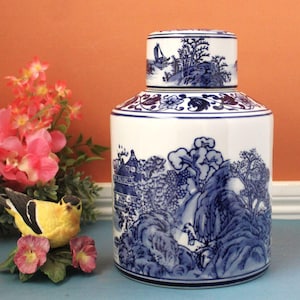 May include: A white ceramic ginger jar with a removable lid, decorated with blue hand-painted illustrations of pagodas, trees, and rocky landscapes. The cylindrical vessel features a floral border pattern around the shoulder.