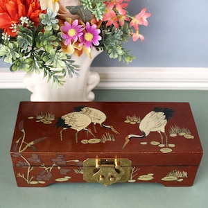 May include: A rectangular wooden box with a dark reddish-brown finish, featuring painted illustrations of white cranes in a marshy landscape. The box includes a brass metal latch on the front and decorative floral motifs along the bottom edge.