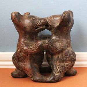 Pode incluir: Uma escultura de argila castanho-escura representando duas figuras animais estilizadas de pé e viradas uma para a outra. As figuras estão unidas pelos braços e apresentam padrões geométricos incisos, incluindo círculos e linhas de grelha, nos corpos.