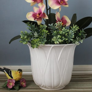 Puede incluir: Una maceta de cerámica blanca con un patrón en relieve de pétalos verticales que contiene una orquídea artificial con flores amarillas y magenta y follaje verde. Una pequeña figura de pájaro amarillo y negro se sitúa junto a la maceta.