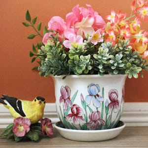 Pode incluir: Um vaso de flores de cerâmica branca com um pires, decorado com íris roxas e azuis pintadas. O vaso é preenchido com folhagem verde e flores rosa e amarelas. Uma estatueta de pássaro amarelo com marcas pretas e brancas está por perto.