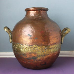 May include: A large, round, hammered copper vessel featuring a wide, textured brass band encircling the middle section. The container has a thick, rolled rim at the top and two curved, metallic handles attached to opposite sides of the body.
