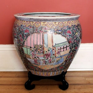 May include: A large, round ceramic fishbowl planter featuring a central cartouche with a detailed scene of figures in traditional attire. The exterior is decorated with a dense floral pattern in pink, green, and blue, resting on a black stand.