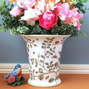 Op de afbeelding: Een witte keramische vaas met een uitlopende bovenkant, versierd met een botanisch ontwerp van wijnranken, bladeren en insecten. De vaas bevat een arrangement van roze en perzik kunstbloemen en groen. Een klein blauw vogelbeeldje staat in de buurt.