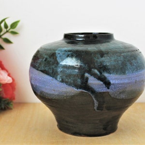 May include: A handcrafted ceramic vase with a rounded body and a dark, glossy finish. The vase features a blend of dark blue, teal, and purple hues, with a dripped glaze effect. The vase has a wide opening and a sturdy base, suitable for displaying flowers or as a decorative piece.