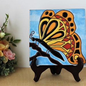 May include: A square ceramic tile featuring a colorful butterfly design. The butterfly has orange, yellow, and red details with black outlines, set against a light blue background. The tile is displayed on a dark brown wooden easel.