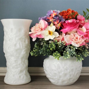 May include: Two white textured vases. One is tall and cylindrical, the other is round and holds a colorful arrangement of pink, orange, purple, and white flowers with green foliage. The vases are set against a blue background.