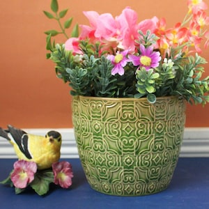 May include: A green ceramic planter with a raised geometric pattern filled with pink and orange artificial flowers. Beside the planter sits a small yellow and black bird figurine perched on a base decorated with two pink flowers and green leaves.