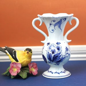 May include: A white ceramic vase with blue floral designs and two curved handles. A yellow and black bird figurine sits next to the vase, perched on a bed of pink flowers and green leaves. The vase is approximately 8 inches tall.