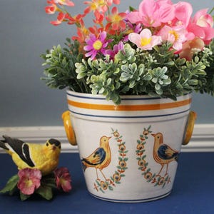 May include: A white ceramic planter with orange and blue stripes and bird illustrations, filled with colorful artificial flowers and greenery. A yellow bird figurine with pink flowers sits nearby. The planter has two yellow handles.