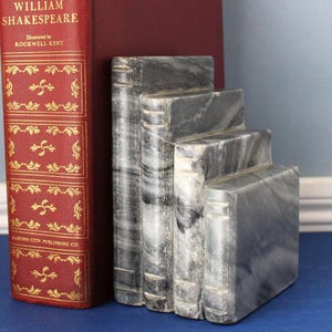 May include: A set of four grey and white marble bookends carved to resemble a stack of books with visible spine details, positioned next to a large red hardcover book titled William Shakespeare illustrated by Rockwell Kent.
