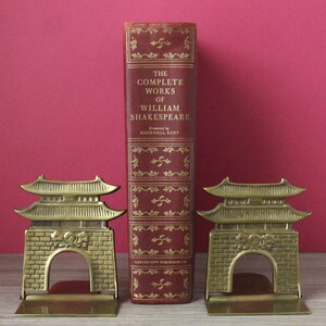 May include: A red hardcover book titled The Complete Works of William Shakespeare with gold lettering and decorative accents, flanked by two brass bookends shaped like traditional East Asian temple gates with brick-patterned bases.