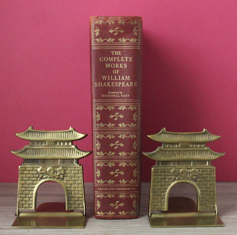 May include: A red hardcover book titled The Complete Works of William Shakespeare with gold lettering and decorative accents, flanked by two brass bookends shaped like traditional East Asian temple gates with brick-patterned bases.