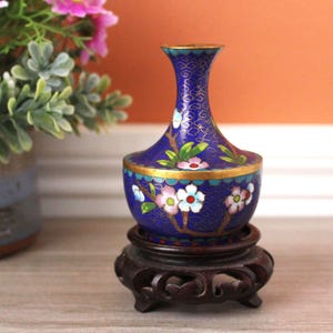 Könnte beinhalten: Eine kleine blaue Cloisonné-Vase mit einem goldfarbenen Drahtmuster und Blumenmotiven in Weiß, Rosa und Grün. Die Vase hat einen schmalen Hals und einen bauchigen Körper und steht auf einem dunkelbraunen, geschnitzten Holzsockel mit filigranen Details.