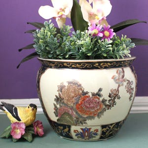 May include: A round ceramic planter with a cream-colored body featuring hand-painted floral motifs in shades of pink, orange, and green, accented by a dark blue rim and base with gold detailing. A small yellow and black bird figurine sits nearby.