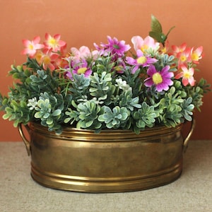 May include: An oval brass planter with two side handles containing a dense arrangement of artificial flowers. The bouquet features small pink, purple, and orange blossoms interspersed with abundant green foliage and small white filler flowers.