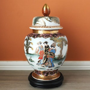 May include: A decorative ceramic ginger jar with a lid, featuring a hand-painted scene of figures in traditional attire. The jar has gold accents and sits on a black wooden base. The lid is topped with a gold finial.