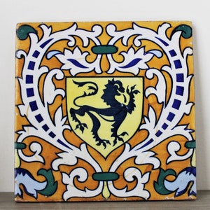 May include: Square ceramic tile with a heraldic design. The tile features a yellow shield with a black lion rampant, surrounded by a decorative border in white, blue, green, and orange. The tile is likely for decorative use.