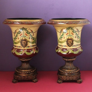 May include: A pair of matching ceramic urns featuring a cream-coloured body with a painted lion head motif, green floral garlands, and red ribbons. The urns have a dark red lower section, a wide flared rim, and a dark bronze-toned pedestal base.
