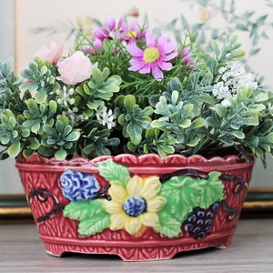 Pode incluir: Um vaso de cerâmica vermelho com uma superfície texturizada, adornado com um girassol, uvas e design de folhas. O vaso é preenchido com folhagem verde, flores rosa e roxas e flores brancas.