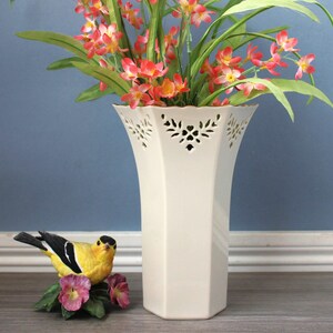 May include: An off-white ceramic vase with a geometric design and decorative cut-outs near the top. It contains red and yellow flowers with green leaves. A small, colourful bird figurine is positioned beside the vase.