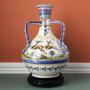 May include: A white ceramic vase with two handles, decorated with a painted scene of two deer in a landscape. The vase has a blue and white striped neck and sits on a black wooden stand. Floral and geometric patterns adorn the base and handles.