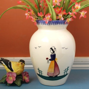 May include: A white ceramic vase with a blue rim and a painted illustration of a woman in traditional clothing holding flowers. The vase contains pink and green artificial flowers. A yellow bird figurine sits nearby.