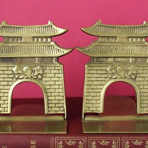 May include: A pair of brass bookends shaped like traditional East Asian gates. Each piece features a multi-tiered roof, a brick-patterned base with a central archway, and a decorative emblem above the arch. The metal has a polished finish.