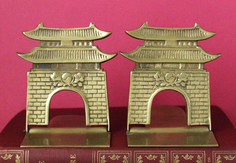 May include: A pair of brass bookends shaped like traditional East Asian gates. Each piece features a multi-tiered roof, a brick-patterned base with a central archway, and a decorative emblem above the arch. The metal has a polished finish.