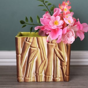 Könnte beinhalten: Ein quadratischer Keramik-Blumentopf mit Bambusdesign in Braun- und Beigetönen. Der Innenraum ist limettengrün. Rosa Blüten mit gelben Zentren und grüne Blätter sind im Inneren des Topfes angeordnet.