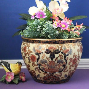May include: A round ceramic planter with a cream-colored base featuring intricate floral patterns in red, blue, and gold, topped with a bouquet of white orchids and pink flowers. A small yellow and black bird figurine sits beside the planter.