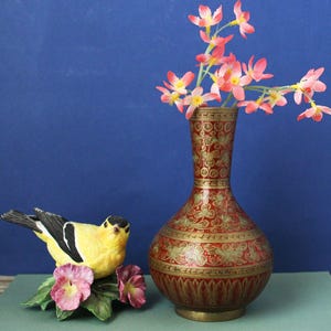 Könnte beinhalten: Eine Messingvase mit roter Emaille-Oberfläche und detaillierten goldfarbenen Blumen- und Blattgravuren, gefüllt mit rosa Blumen. Daneben steht eine gelb-schwarze Vogelfigur, die auf einem Sockel aus grünen Blättern und rosa Blüten ruht.