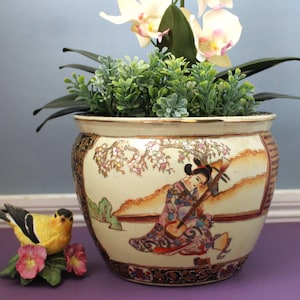 May include: A ceramic planter with a cream-colored base featuring a painted scene of a person playing a stringed instrument under a flowering tree. The rim is gold-toned, and the base has a dark, intricate border. A small yellow bird figurine sits nearby.