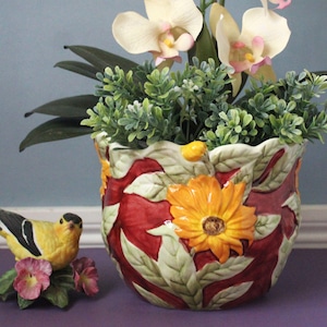May include: A ceramic planter featuring a deep red background with raised, three-dimensional yellow flowers and green leaves. Beside the planter sits a small, yellow and black bird figurine perched on a cluster of pink flowers and green leaves.