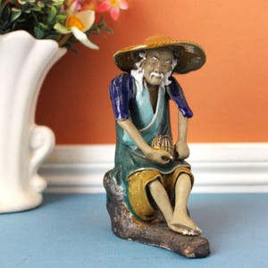 May include: A ceramic figurine of an elderly person wearing a wide-brimmed straw hat, a blue and teal tunic, and mustard yellow trousers. The figure is seated on a dark, textured rock-like base, holding a small, round, brown-striped object.