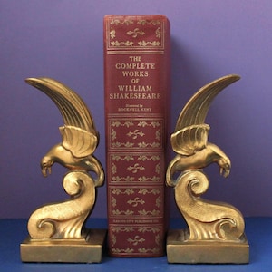 May include: A red hardcover book titled The Complete Works of William Shakespeare illustrated by Rockwell Kent, flanked by two gold-colored metal bookends shaped like birds perched atop stylized swirling wave patterns on square bases.