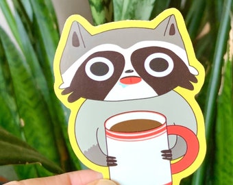 May include: A cartoon raccoon sticker with a cute expression, holding a red and white coffee mug with brown coffee inside. The sticker has a yellow outline.