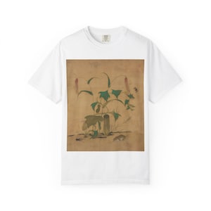 以下が含まれることがあります： ヴィンテージの植物画が描かれた白いTシャツ。緑の葉、ピンクの花、茶色の背景が特徴です。シャツは、クラシックなクルーネックと半袖です。