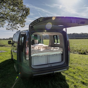 May include: A dark-colored van with its rear hatch open, revealing a sleeping area inside. The interior features a mattress with light-colored bedding placed on top of two grey plastic storage crates, creating a compact mobile living space.