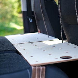 Könnte beinhalten: Ein weißer Holztisch mit einem Loch in der Ecke, befestigt an der Rückseite von zwei schwarzen Autositzen. Der Tisch ist aus Sperrholz und hat ein Gitter aus kleinen Löchern.