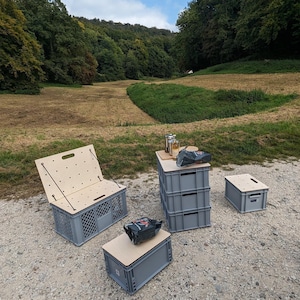 Könnte beinhalten: Ein Set aus grauen Kunststoff-Aufbewahrungsboxen mit hellen Holzdeckeln, bestehend aus einer Box mit hochgeklapptem Deckel, einem Stapel aus drei Boxen, einer einzelnen flachen Box und einer kleinen Box, angeordnet auf einem Kiesweg im Freien.