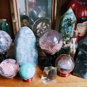May include: A collection of polished crystals and gemstones in various shapes and colors. Spheres, obelisks, and freeform stones are displayed on a wooden shelf. The crystals include shades of purple, green, clear, and brown, with some resting on decorative stands.