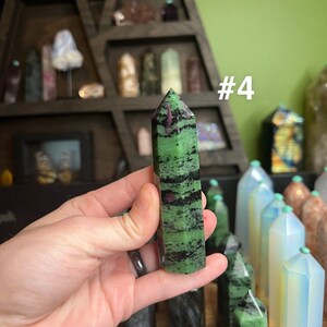May include: A green and black striped crystal point with a pink stripe, labeled #4. The crystal is being held in a hand.
