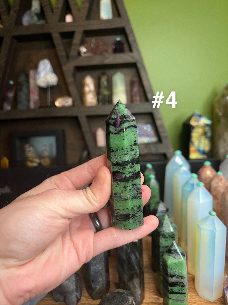May include: A green and black striped crystal point with a pink stripe, labeled #4. The crystal is being held in a hand.