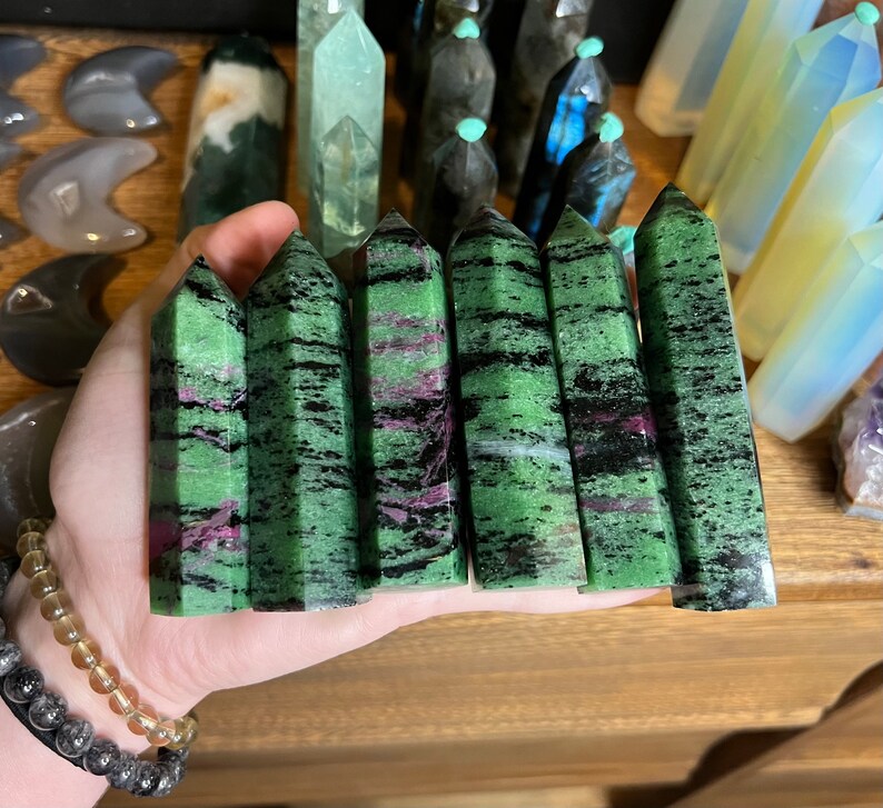 May include: Seven green, black, and pink gemstone points, also known as green aventurine, are held in a hand. The points are polished and have a smooth, shiny finish.