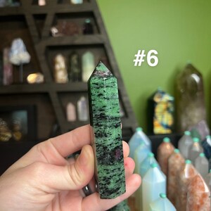 May include: A green and black zultanite point with a pink hue. The point is about 2 inches long and is held in a hand. The point is labeled #6.